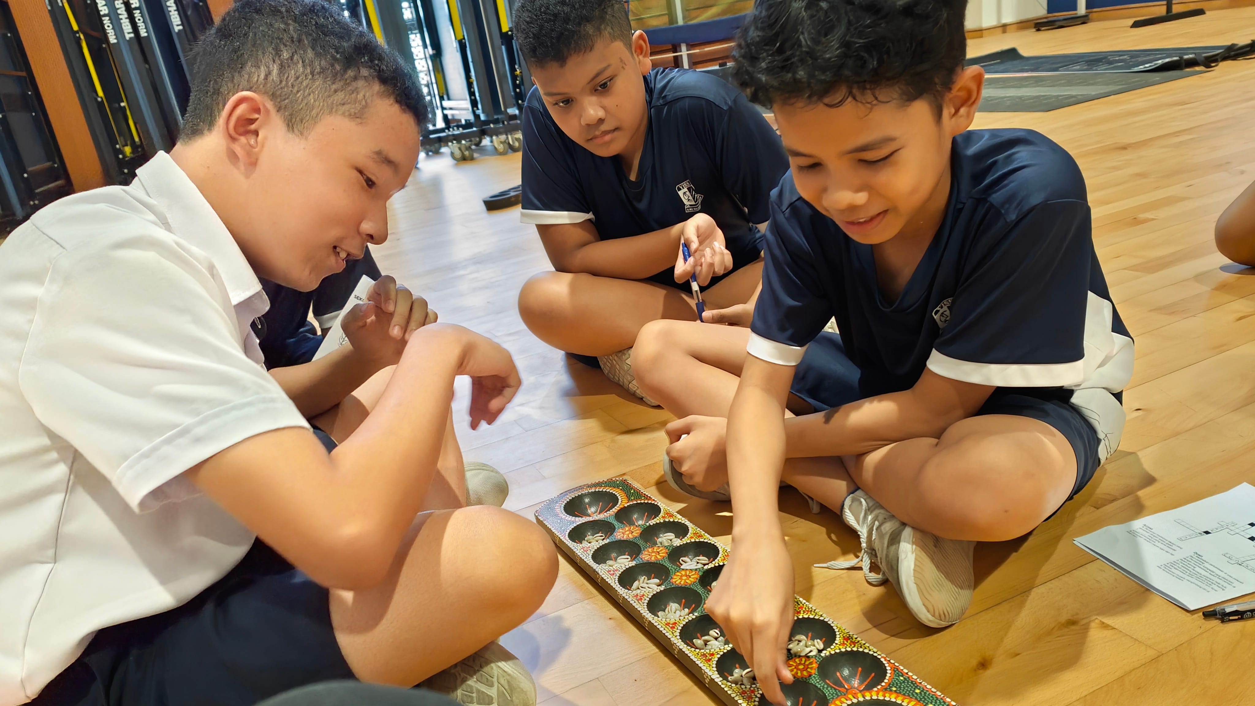 Three boys in school uniforms sit on a wooden floor playing a mancala board game with shells.